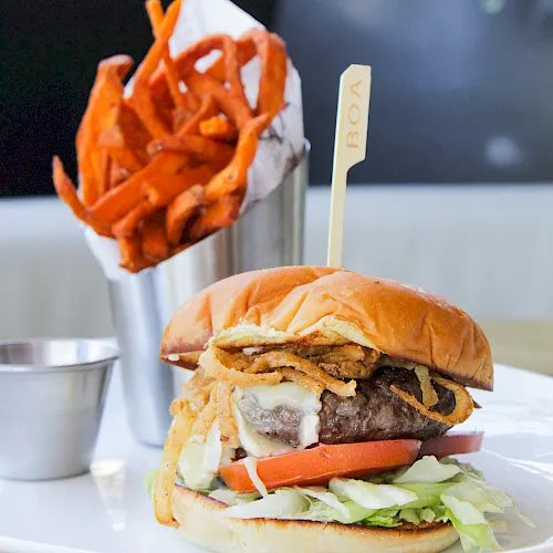 A juicy burger with melted cheese and veggies sits on a plate beside a cup of crispy orange fries, ready to eat.