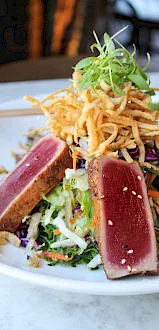 A plate of mixed greens and shredded veggies topped with crispy noodles and two seared tuna steaks.