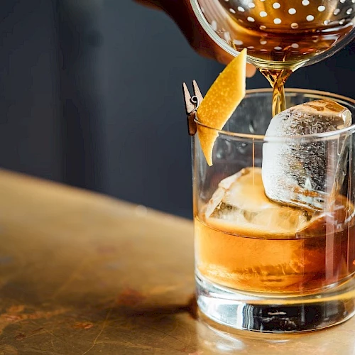 A cocktail glass with a carved ice cube, orange twist, and a black-and-gold spoon pouring liquor into a lowball glass, set on a wooden bar top.