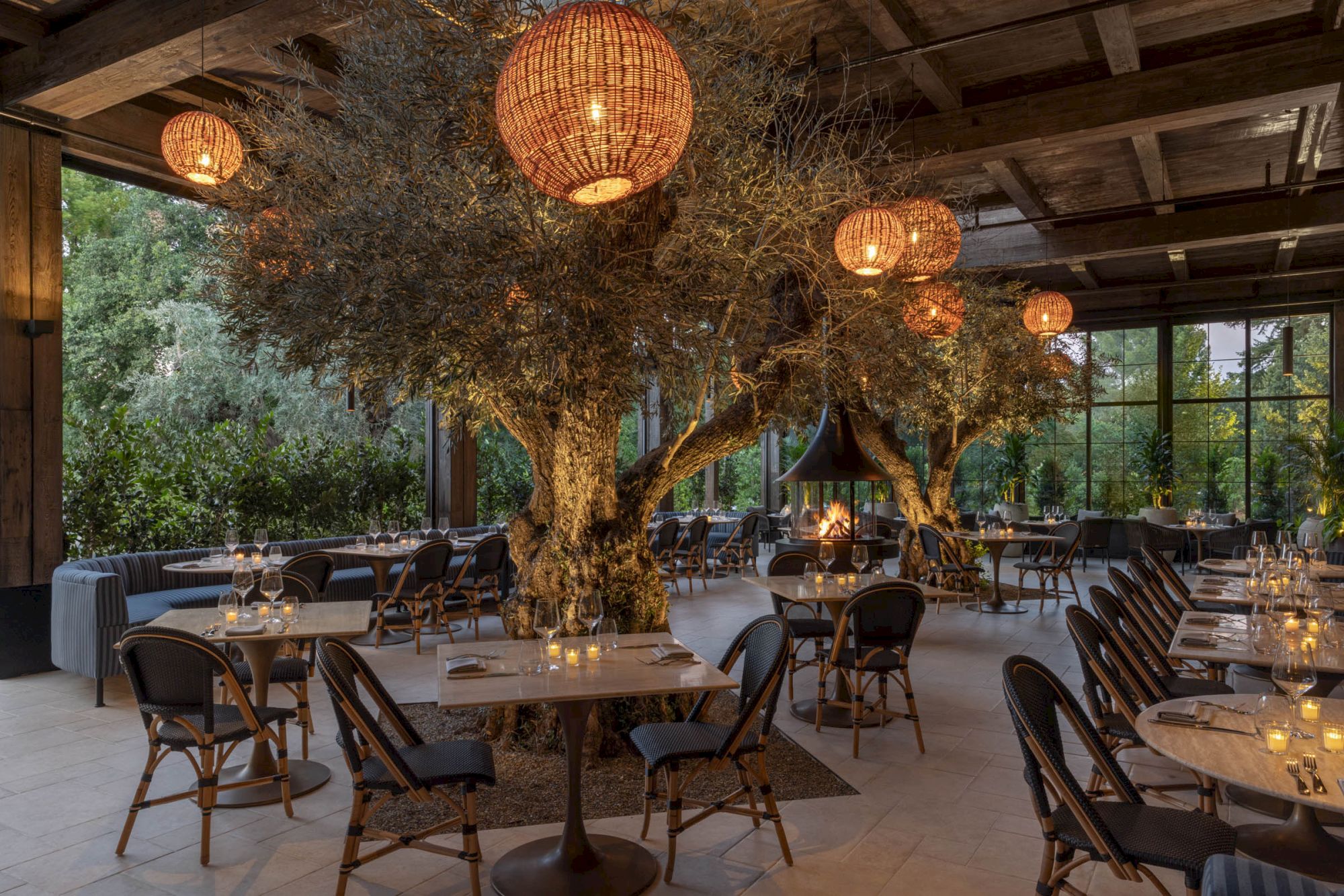 An elegant indoor dining area with candlelit tables, hanging woven lanterns, and giant faux trees around a cozy fire pit.