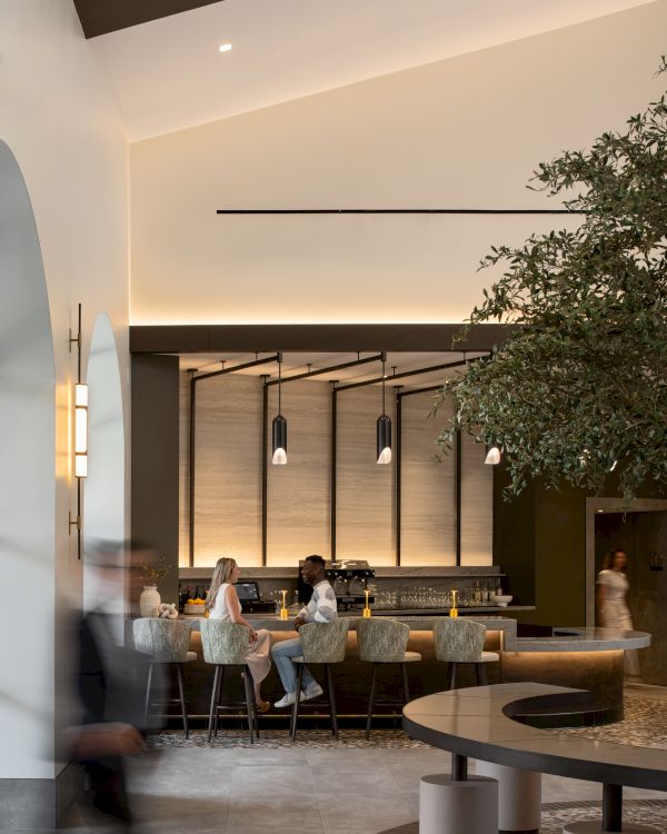 A modern hotel lobby with a long bar, warm lighting, trees indoors, and people seated at the bar while others walk by, airy and stylish atmosphere.