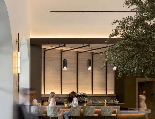 A modern hotel lobby with a long bar, warm lighting, trees indoors, and people seated at the bar while others walk by, airy and stylish atmosphere.
