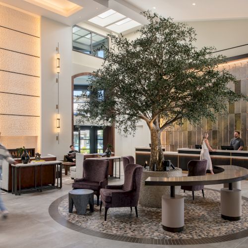 A modern hotel lobby with a large indoor tree centerpiece, stylish seating, a front desk, and guests checking in, creating a calm, upscale vibe.