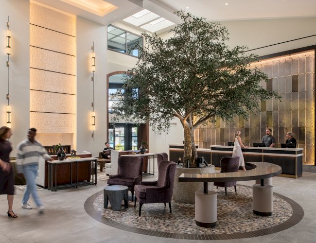 A modern hotel lobby with a large indoor tree centerpiece, stylish seating, a front desk, and guests checking in, creating a calm, upscale vibe.