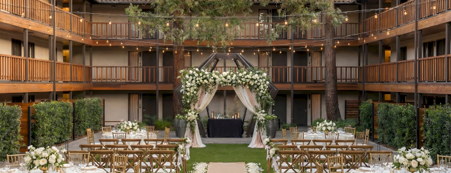A European-style courtyard with a central lawn, surrounded by two-story brick buildings, stone arches, and white-clothed tables set for outdoor dining.