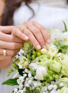 A couple holds a white bridal bouquet together, with the bride's and groom's hands displaying rings over white flowers and greenery.