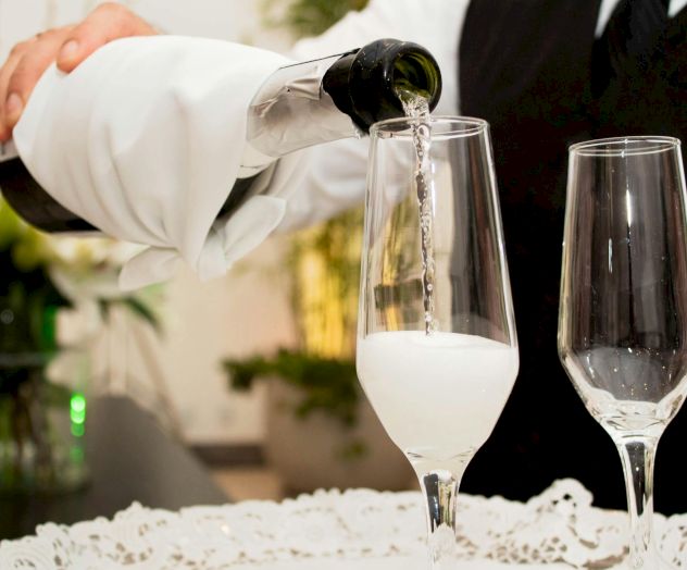 A person pours white wine into a flute, with another empty glass beside it, at a crafted table setting.