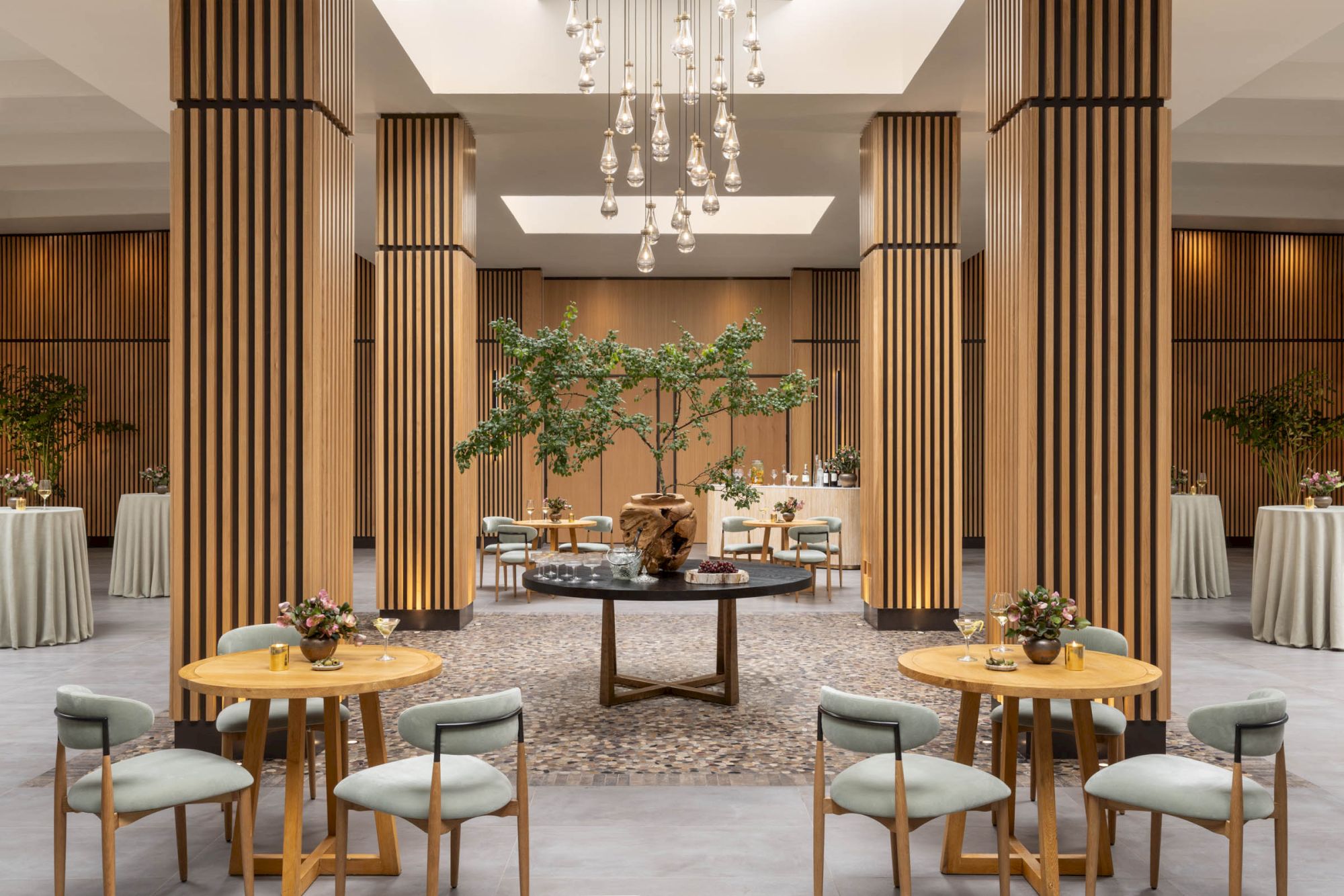Elegant event dining room with wooden pillars, glass chandeliers, round tables, and flowers in a central lounge area.