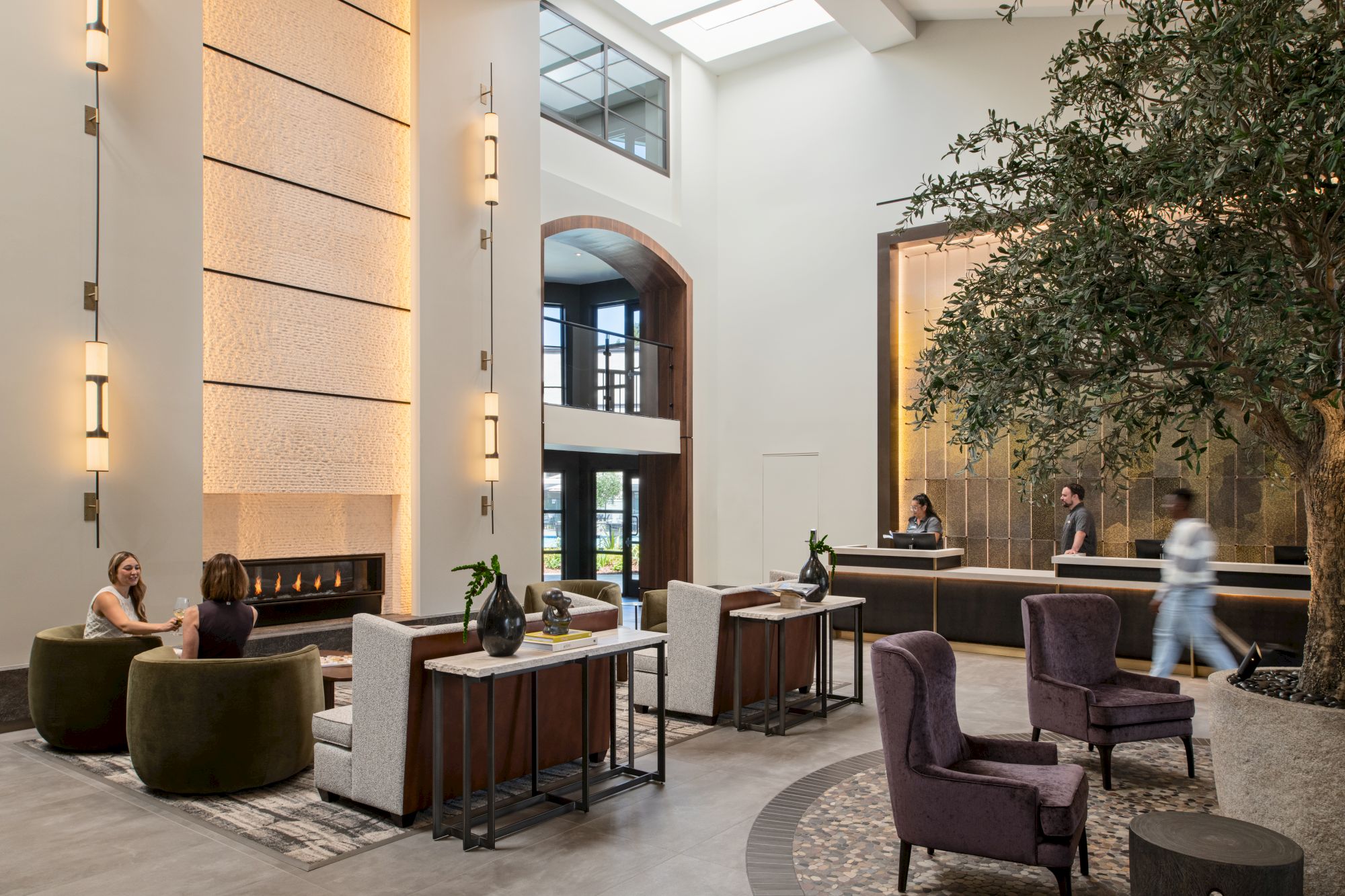 A modern hotel lobby with high ceilings, tall lights, seating areas, and a reception desk; people mingle near lush trees and stylish furniture.