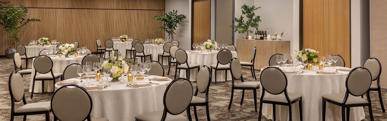 A well-lit event hall set for a formal dinner, with round tables, chairs, candles, glassware, and floral centerpieces.