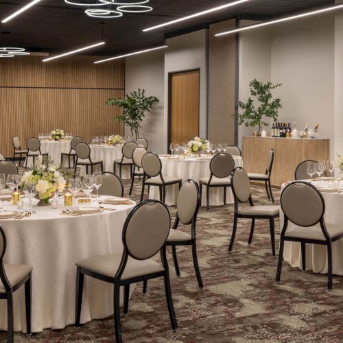 A well-lit event hall set for a formal dinner, with round tables, chairs, candles, glassware, and floral centerpieces.