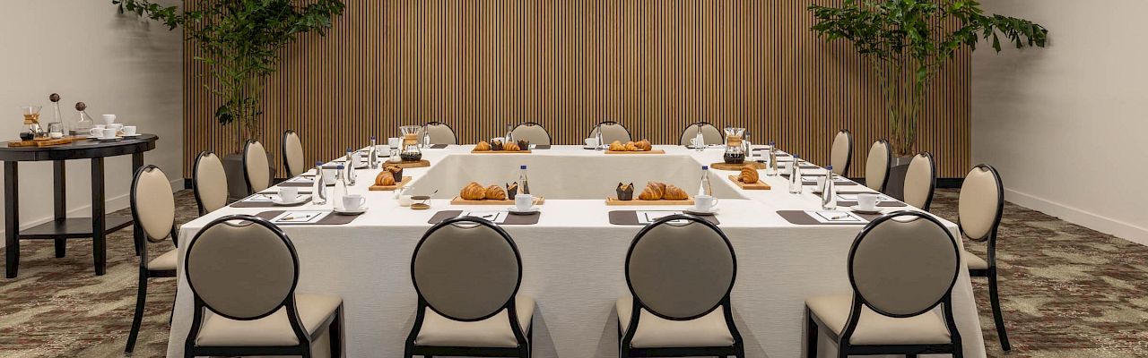 A modern meeting room set with a long banquet table, chairs, pastries, and green plants under LED ceiling lights.