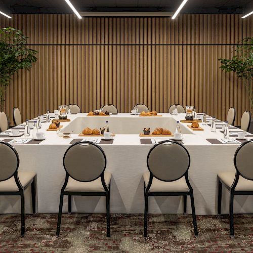 A modern meeting room set with a long banquet table, chairs, pastries, and green plants under LED ceiling lights.