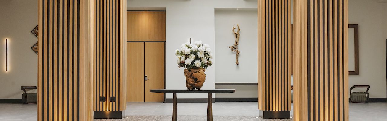 A spacious, modern lobby with tall wooden slatted columns, a central vase of white flowers on a dark table, and soft, ambient lighting.