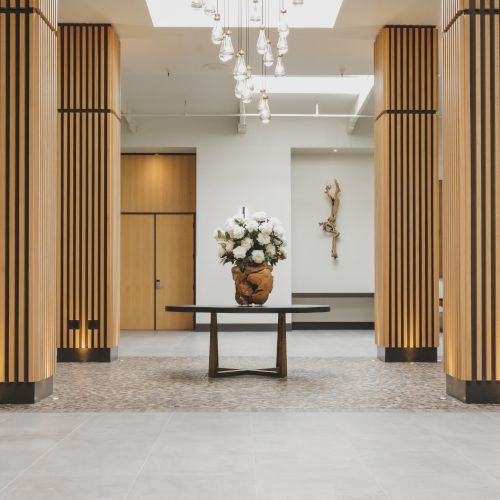 A spacious, modern lobby with tall wooden slatted columns, a central vase of white flowers on a dark table, and soft, ambient lighting.