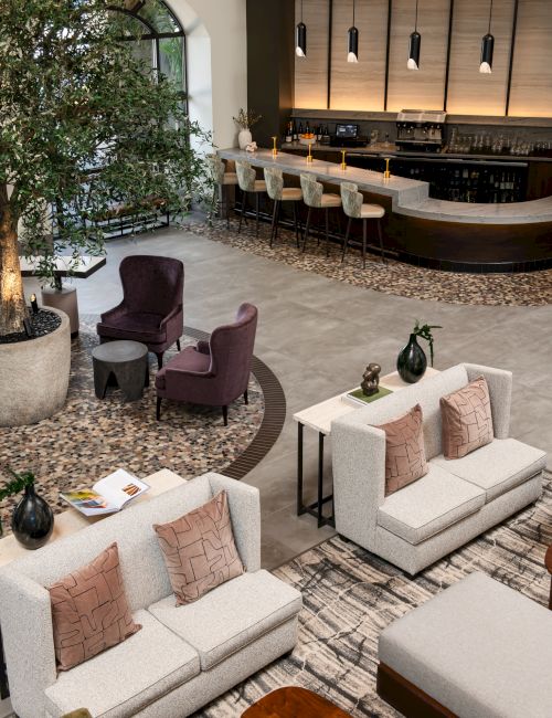 A modern hotel lobby with a curved bar, warm lighting, tall bar stools, beige sofas, neutral cushions, potted trees, and a cozy seating area.