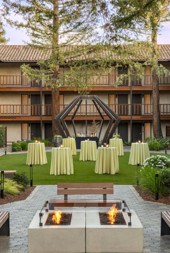 A hotel courtyard with a central fire pit, tall trees, patio seating, and round alta tables with yellow tablecloths, surrounded by a U-shaped building.