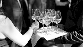 A server offers a tray of wine glasses to a guest at a formal event, with blurred guests in the background.
