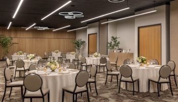 A modern banquet hall set with round tables, candles, glassware, flowers, and sleek ring chandeliers under bright ceiling lights.
