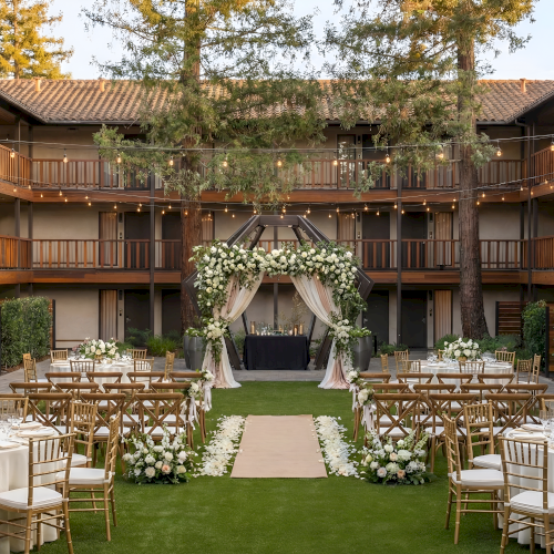 Outdoor wedding setup in a courtyard with a floral arch, wooden chairs, round tables, and elegant centerpieces on green lawn. Ends right.