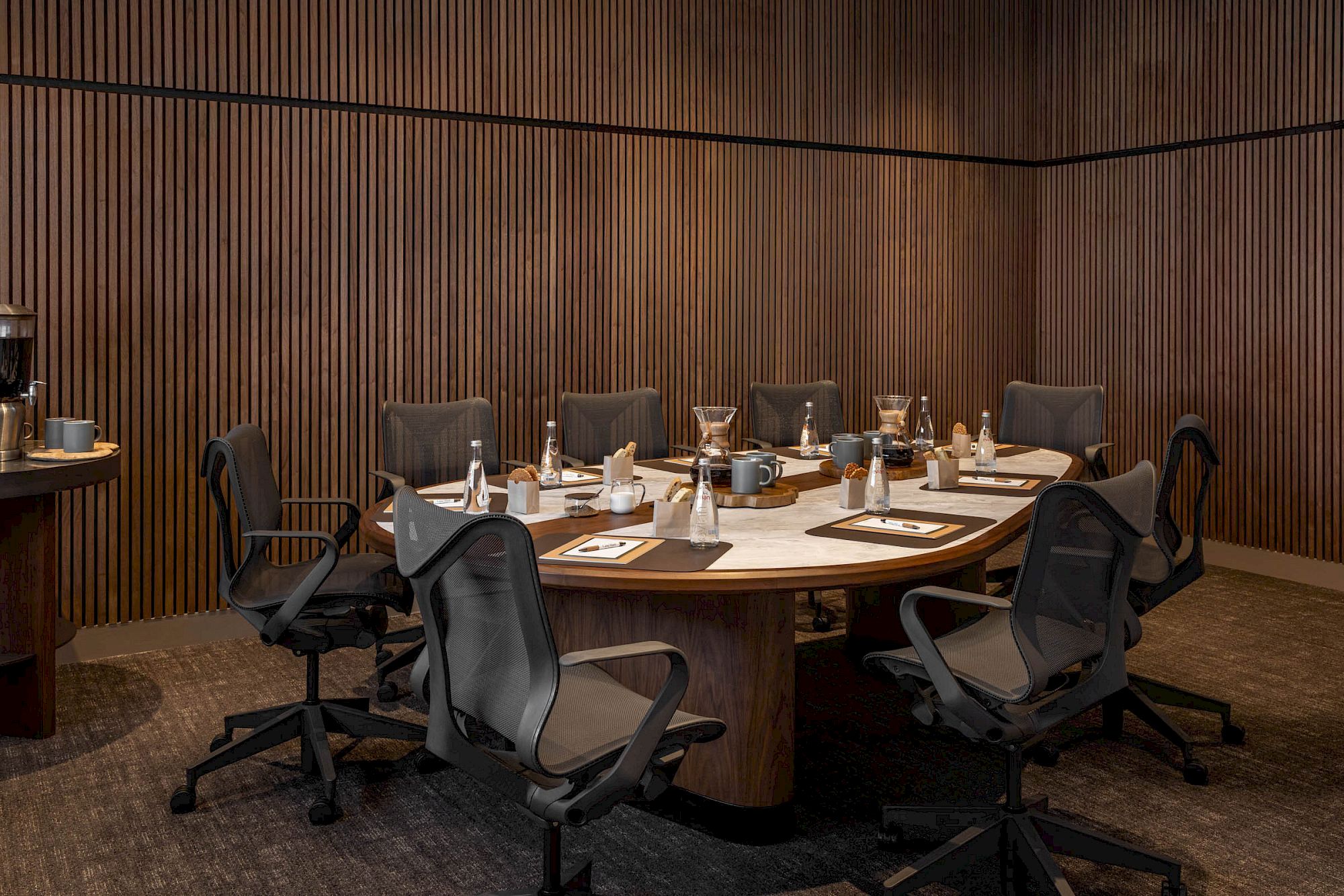 A modern meeting room with a round table, office chairs, and coffee/serving items arranged on place settings, ready for a discussion.