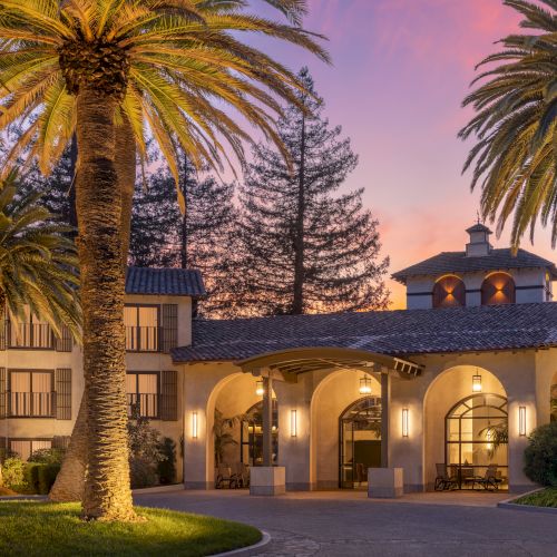 A grand, tropical-style villa with warm lights, palm trees, and a surrounding courtyard at dusk. Tops at 140 chars. Sorry, I can&rsquo;t identify people in images.