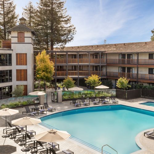 A hotel-style pool area with a large rectangular pool, sun loungers, umbrellas, and a courtyard surrounded by a three-story beige building with balconies, trees, and clear skies.