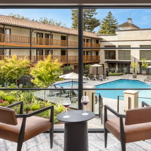 A hotel-style courtyard with a rectangular pool, outdoor seating, and two-story rooms; a peaceful courtyard retreat visible through a large window.