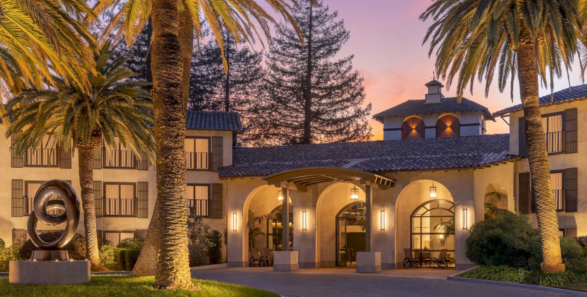 A sunny resort courtyard at sunset with tall palm trees, warmly lit stucco arches, tiled roofs, and a circular metal sculpture in the garden.