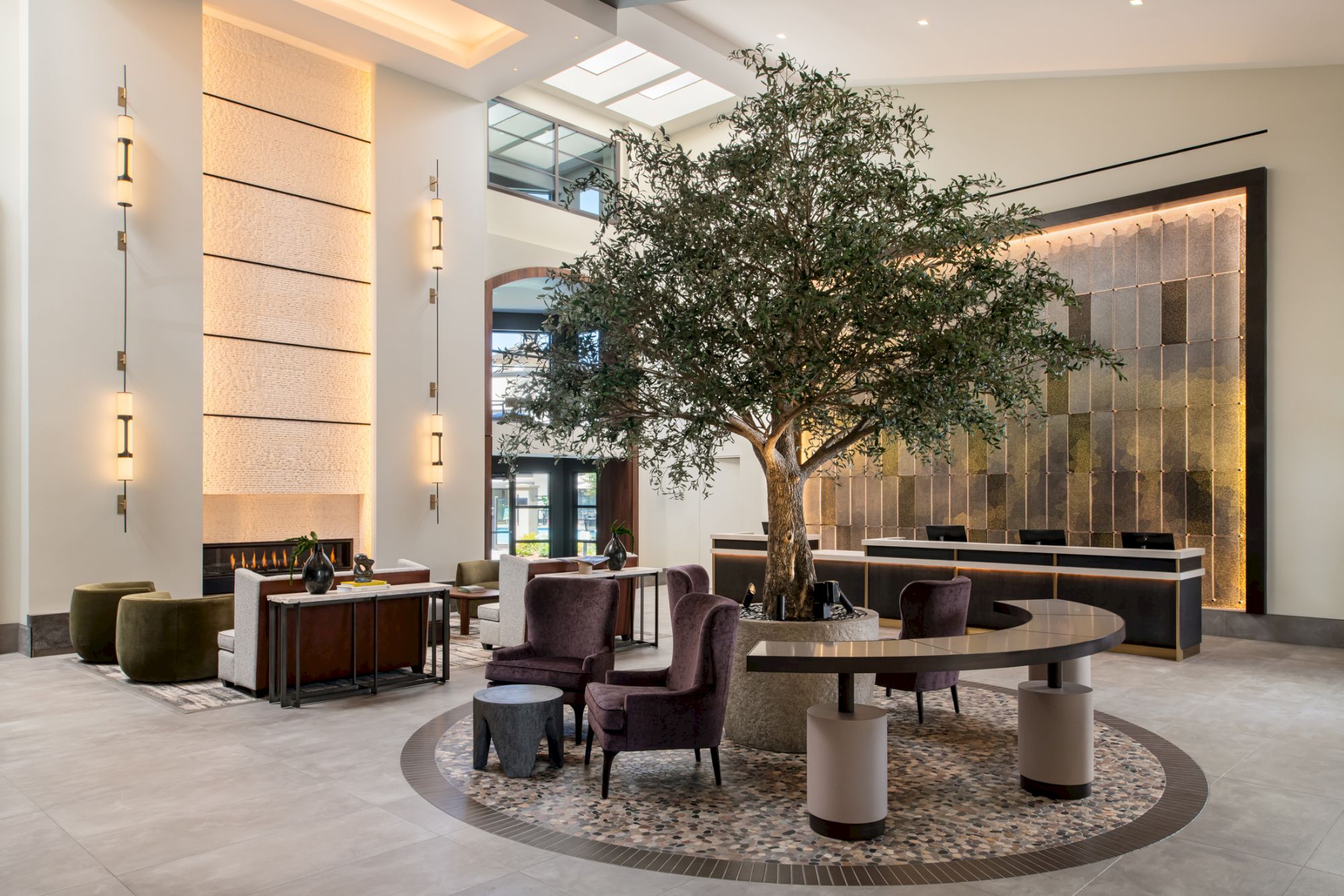 A modern hotel lobby with a large tree centerpiece, stylish seating around circular rug, neutral tones, tall wall lights, and a decorative wall panel; elegant and spacious ambiance.