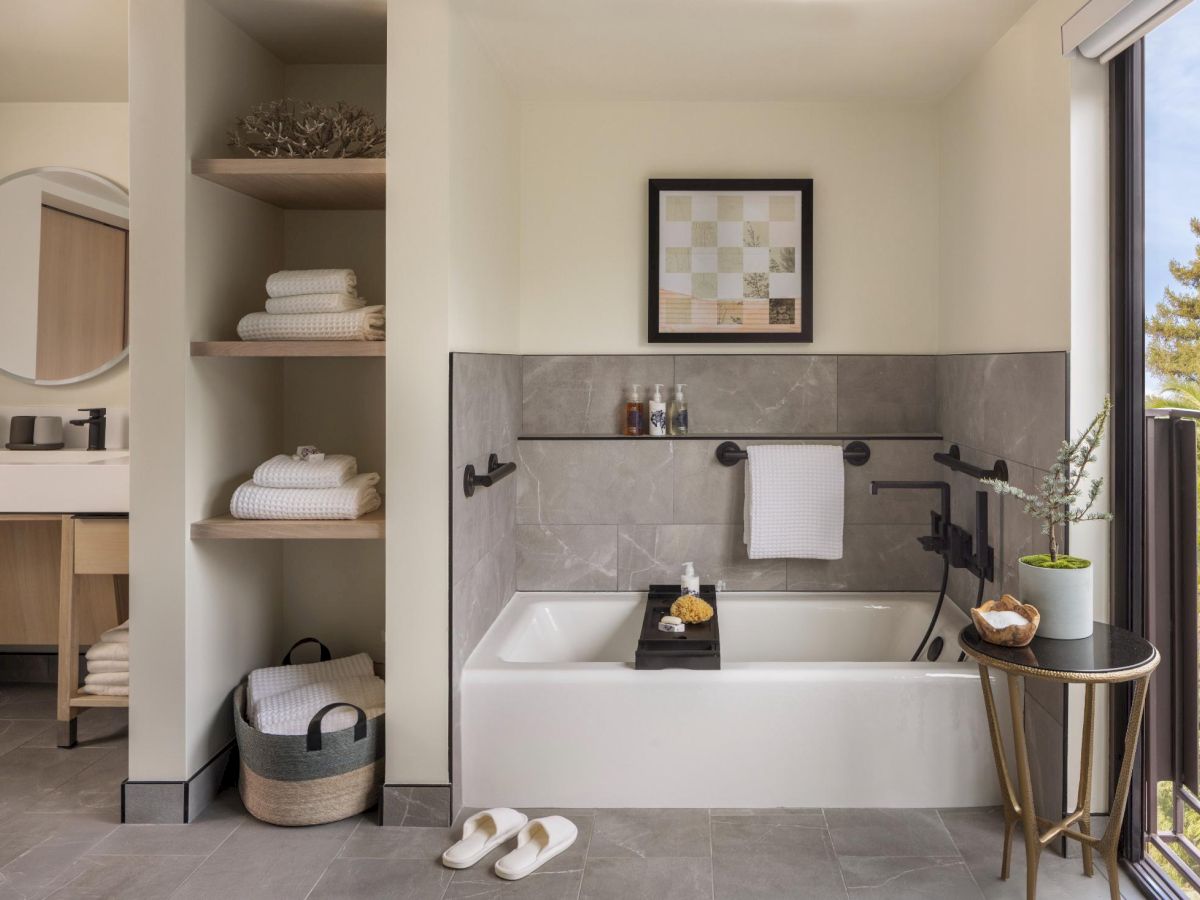 A modern hotel bathroom with a bathtub, open shelving, a balcony view, and neatly arranged towels and decor, all in light gray tones.