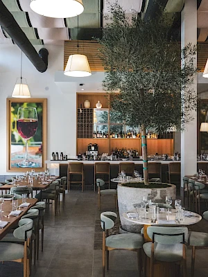 Cozy, modern dining space with round marble tables, soft upholstered chairs, warm lighting, a central tree, and a bar area in the back.