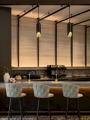 A modern bar or cafe interior with a long counter, three upholstered stools, pendant lights, a backdrop of panels, and a tiled floor.