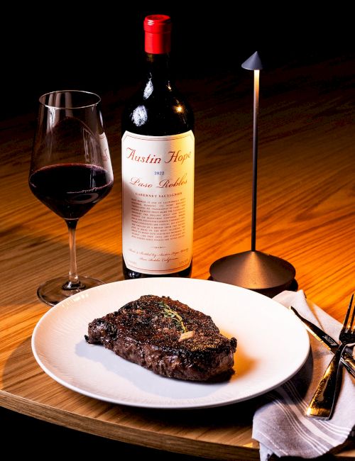 A dining set with a steak on a plate, a red wine bottle and glass, a candle, and fork and knife on a wooden table.