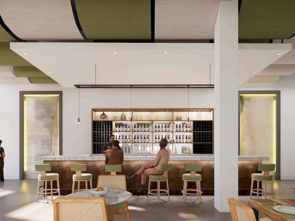 A modern bar or cafe with a long wooden counter, bar stools, shelves of bottles, and a few patrons seated and standing, bright and airy interior.