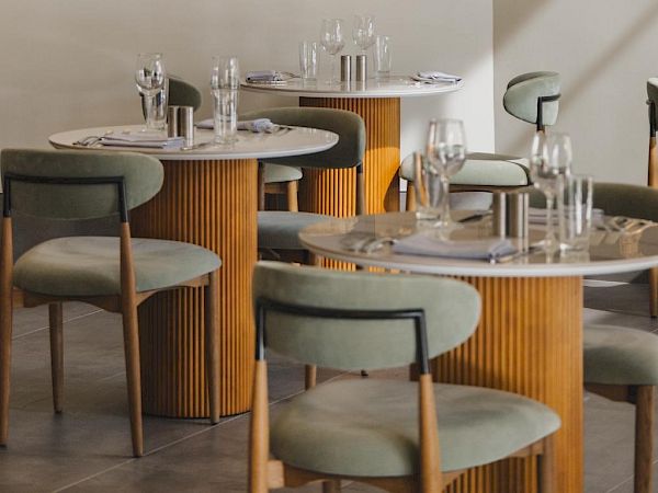 A stylish restaurant interior with round marble-topped tables, light wood pedestal bases, and soft green upholstered chairs.
