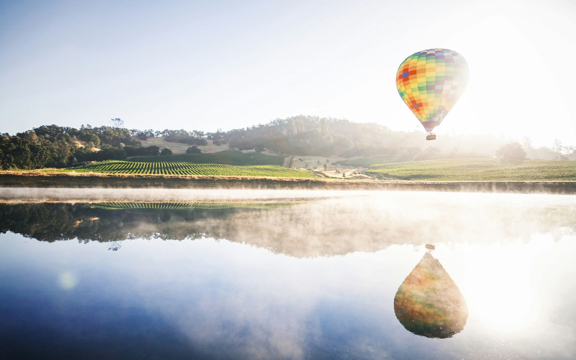 A colorful hot air balloon floats above a calm lake, reflecting perfectly on the water as hills and fields stretch into the distance.
