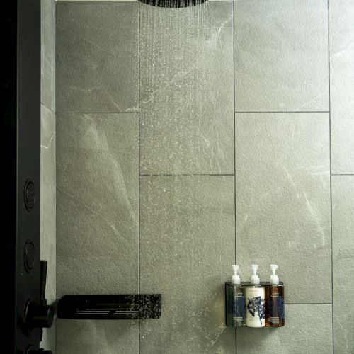 A modern shower with a large black rain showerhead, dark fixtures, and a small shelf holding three pump bottles on gray tiled walls.