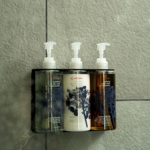 Three bottles of liquid soap or lotion mounted on a small wall shelf with pumps, against a tiled wall.