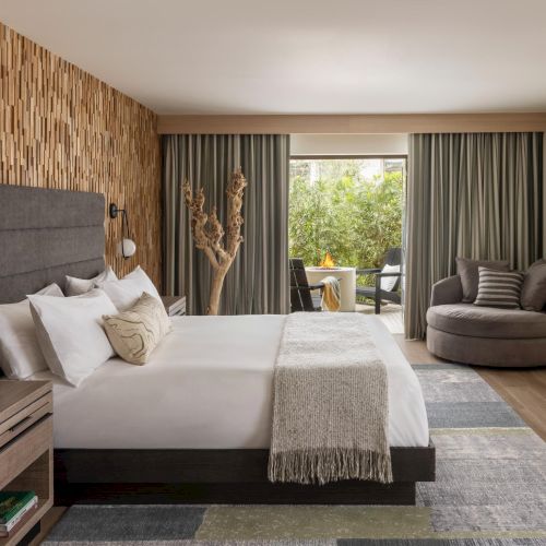 A bright, stylish bedroom with a large bed, built-in headboard, soft lighting, a seating area, and greenery visible through sliding glass doors, inviting and serene.