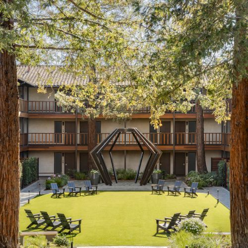 A courtyard with a manicured lawn, surrounded by a U-shaped wooden apartment building, tall trees, and several lounge chairs set up around the grass.