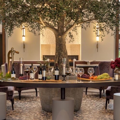 A group of people sits around a long dining table in a bright, airy room with large windows and plants; warm, casual gathering near sunset.