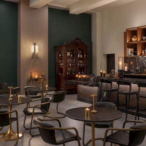 An elegant candlelit bar lounge with marble countertop, liquor bottles, armchairs, and stools in warm, moody lighting.