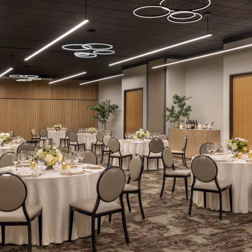 A modern banquet hall set for a wedding or event, with round tables, chairs, candles, flowers, and elegant ring ceiling lights.