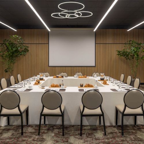A modern meeting room with a long set table for catering, chairs, potted trees, and a large ceiling light above.