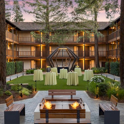 A serene indoor conservatory with a glass ceiling, lush plants, and a long wooden table set for dining, bathed in natural light.