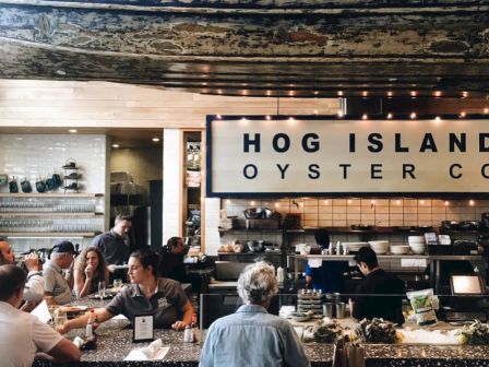 A bustling seafood restaurant with diners at tables, a chef area, and a large sign reading &ldquo;HOG ISLAND OYSTER CO&rdquo; in a bright, modern interior.