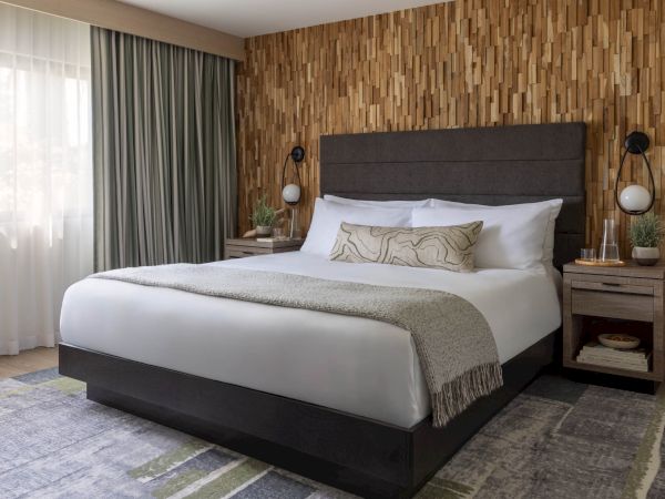 A modern bedroom with a large bed, patterned pillows, bedside lamps, and wood wall paneling.