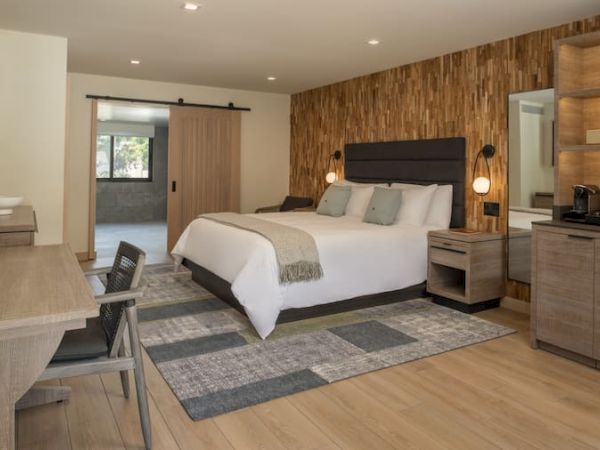 A modern hotel room with a king bed, wood accent wall, nightstands, a dresser, a small desk, and a kitchenette area, all in neutral tones.