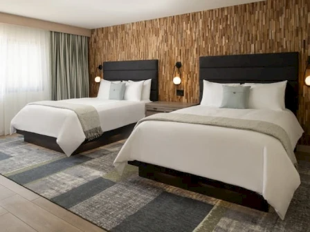 Two neatly made hotel beds in a modern room with a wood accent wall, soft lighting, and a patterned rug, perfect for a restful stay.